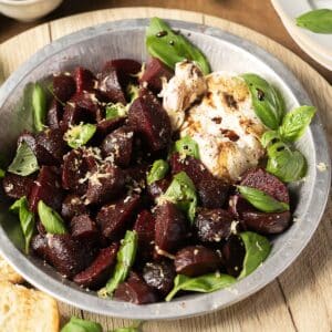 A metal dish with roasted beet cubes, fresh basil leaves, a dollop of burrata cheese, lemon zest, and a drizzle of balsamic glaze.