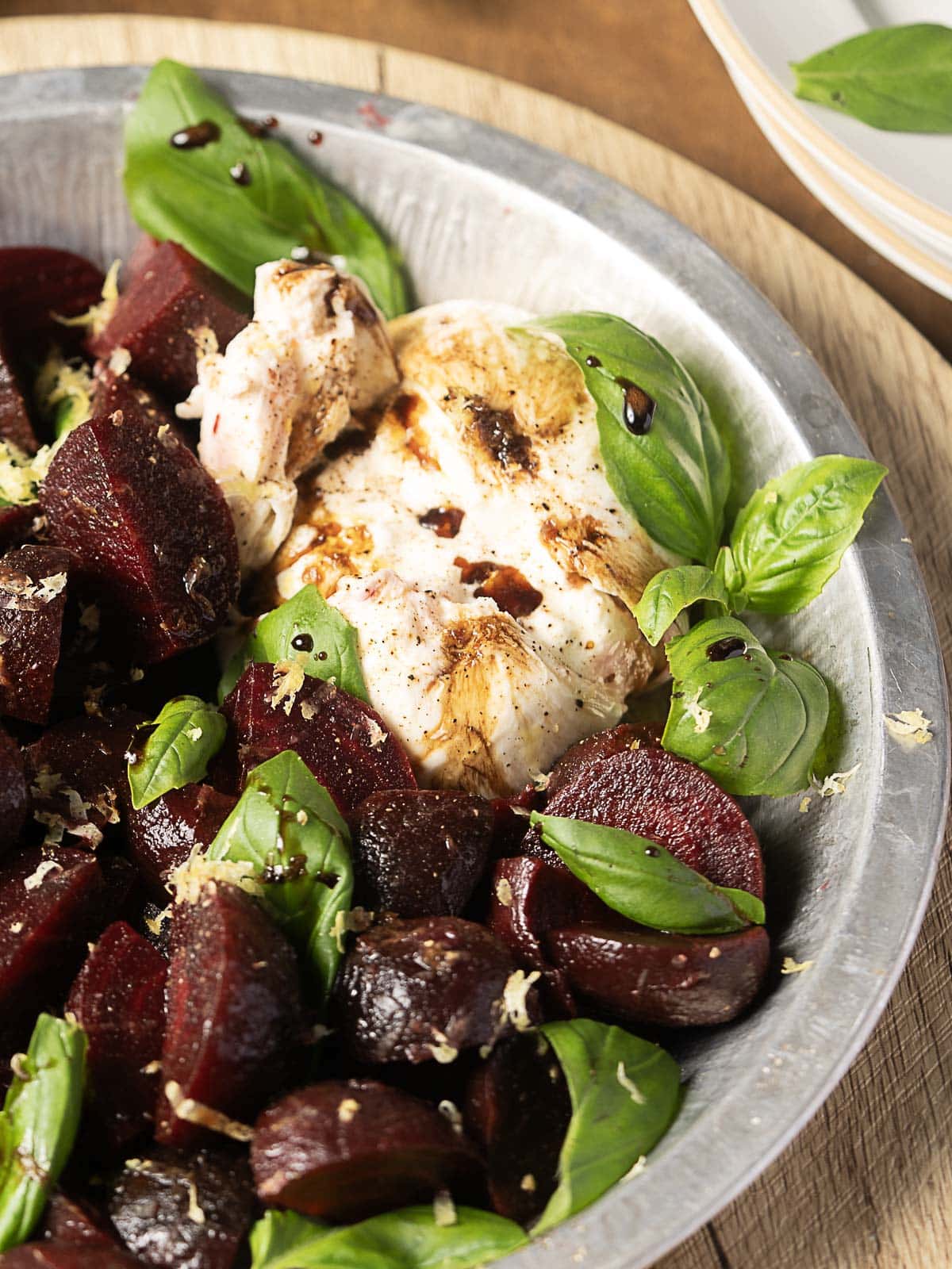 A metal bowl filled with cooked beetroot chunks, fresh basil leaves, a portion of burrata cheese, grated lemon zest, and a drizzle of balsamic glaze.