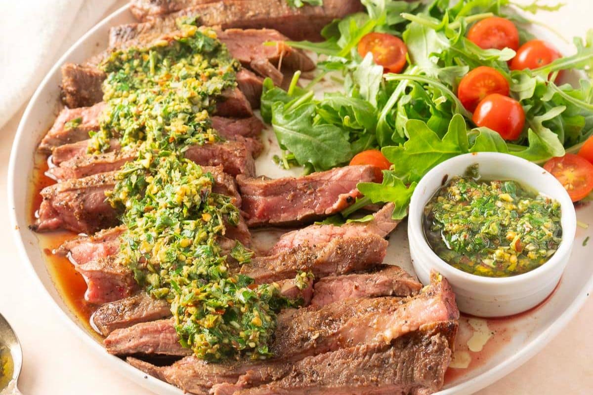 Sliced steak topped with chimichurri sauce, served with a side of arugula and cherry tomatoes, and extra chimichurri in a small bowl.