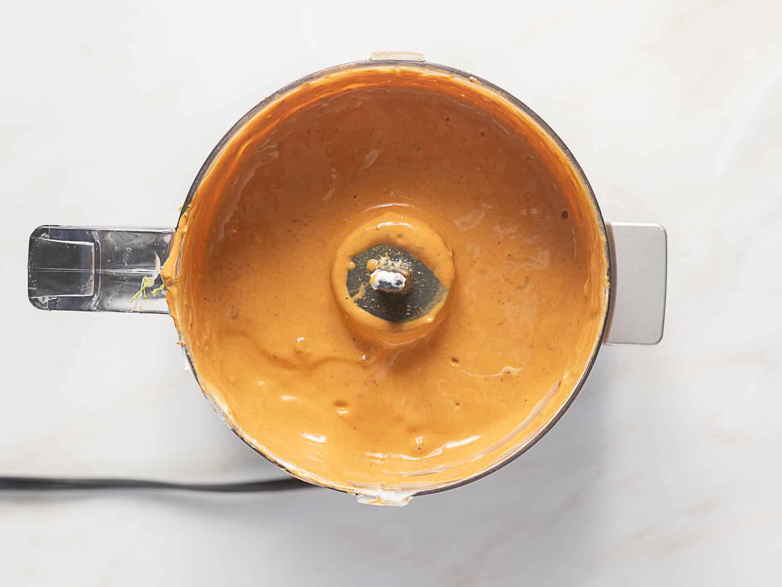Overhead view of a food processor containing smooth, orange-red sauce or puree with a visible metal blade in the center.