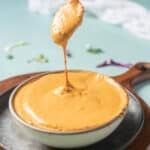 A spoon lifts creamy, orange-colored sauce from a bowl placed on a round tray, with a blue background and some scattered herbs.
