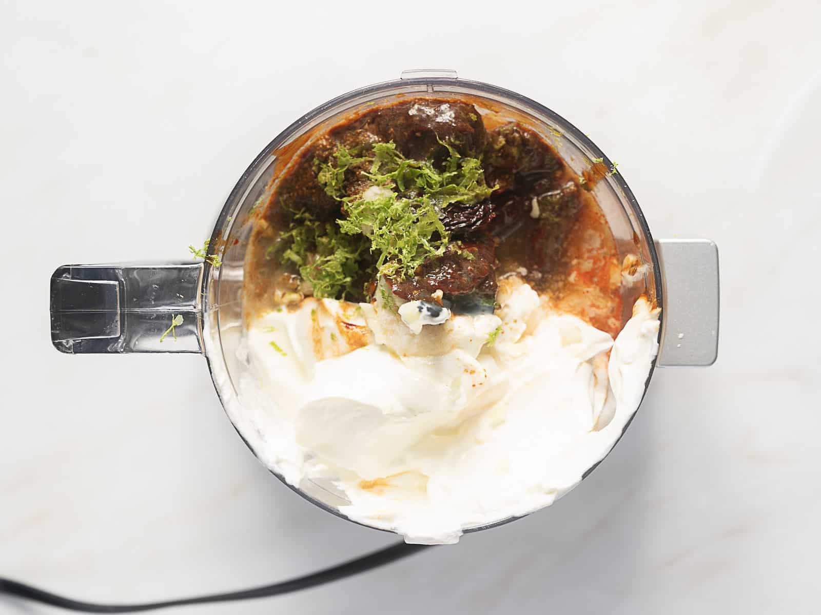 Top view of a food processor containing yogurt, spices, herbs, and sauce ingredients ready to be blended.