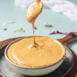 A spoon lifts creamy, orange-colored sauce from a bowl placed on a round tray, with a blue background and some scattered herbs.