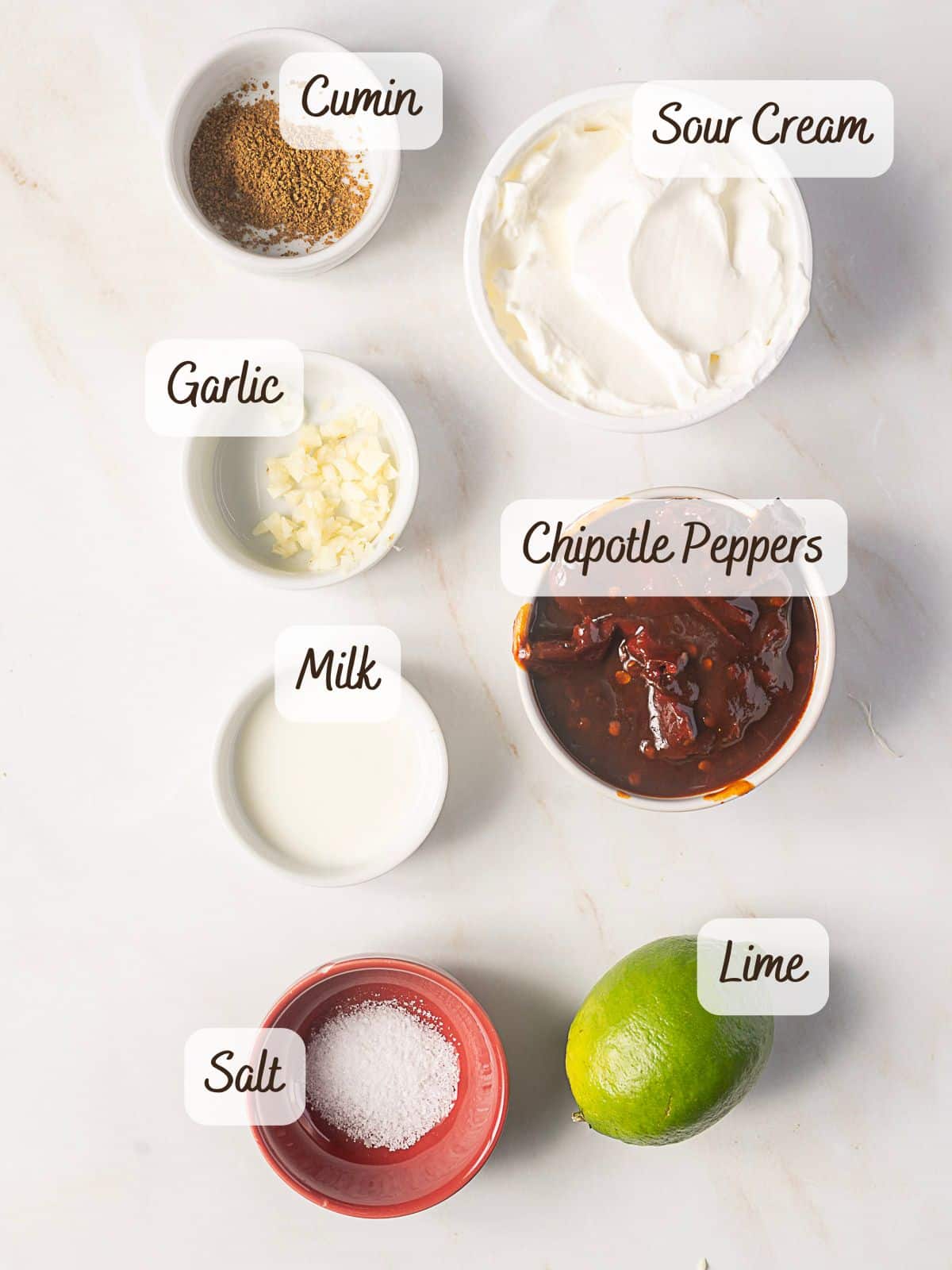 Bowls of cumin, sour cream, chopped garlic, chipotle peppers, milk, salt, and a whole lime arranged on a white surface—each labeled with text—perfect ingredients for making homemade chipotle sauce.