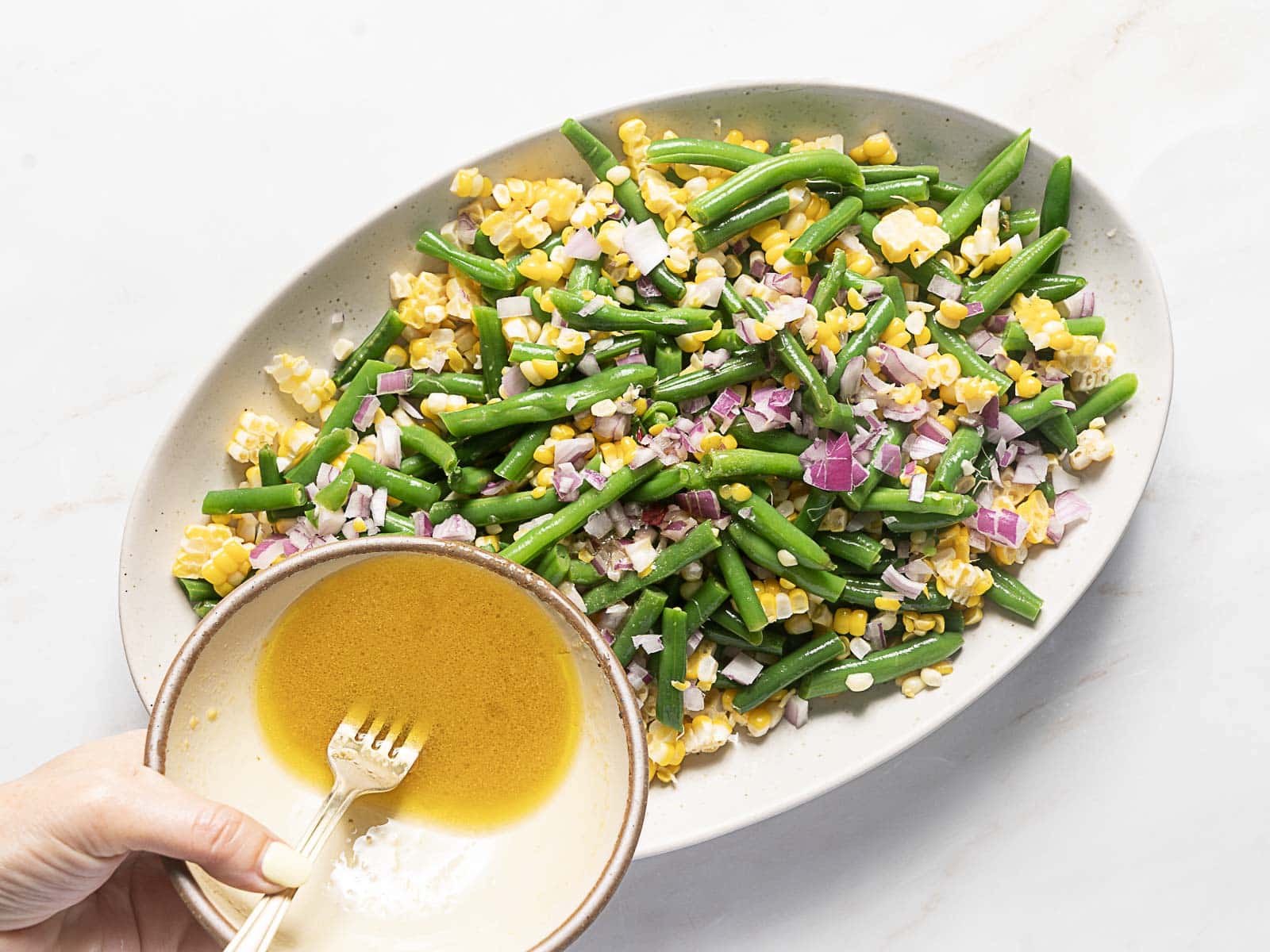 A hand holding a bowl of dressing above a platter of green beans, corn, and chopped red onions, ready to be poured over the vegetables.