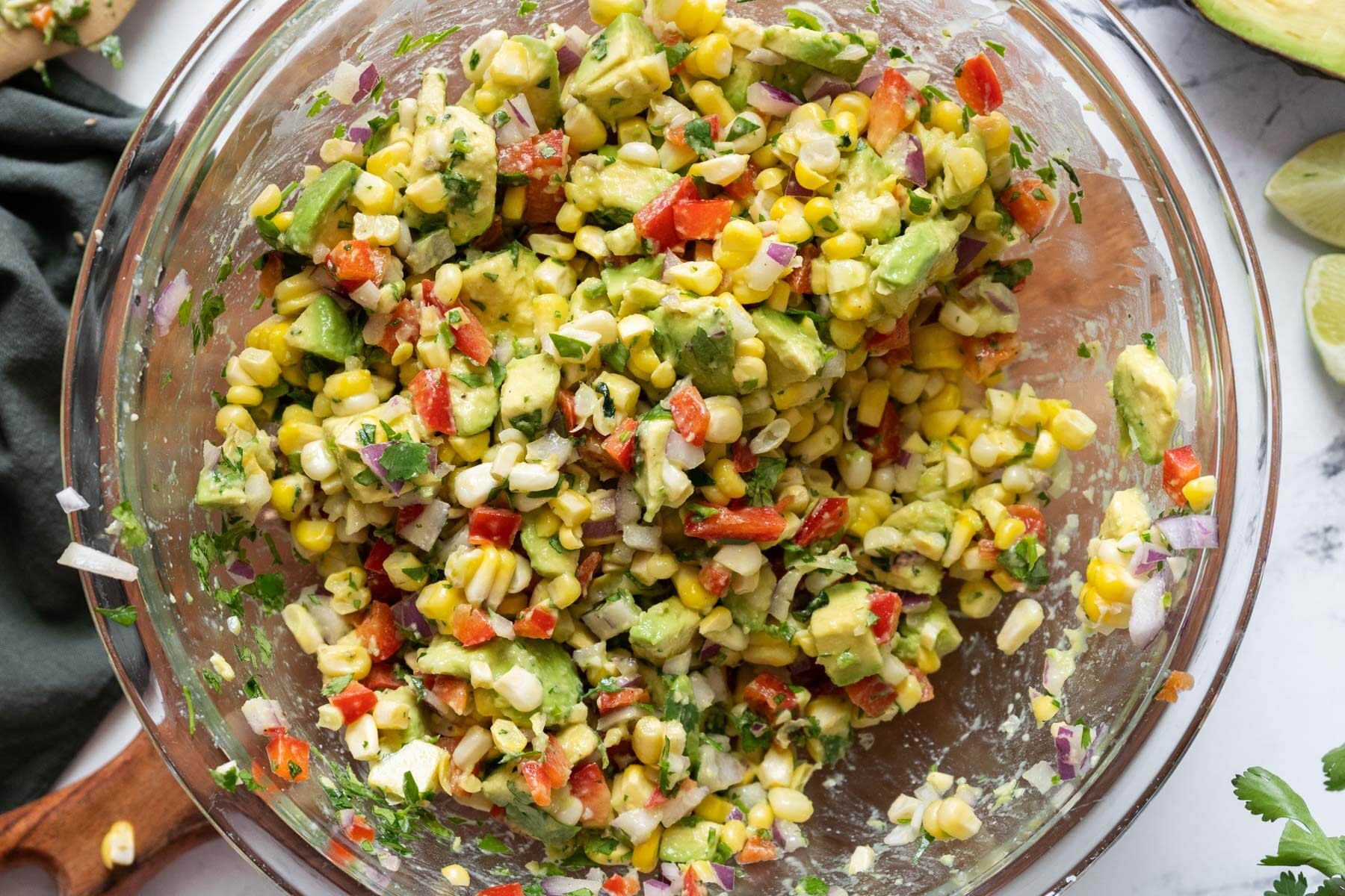 A glass bowl filled with a colorful mixture of corn, avocado, red bell pepper, red onion, and herbs, sitting on a marble surface.