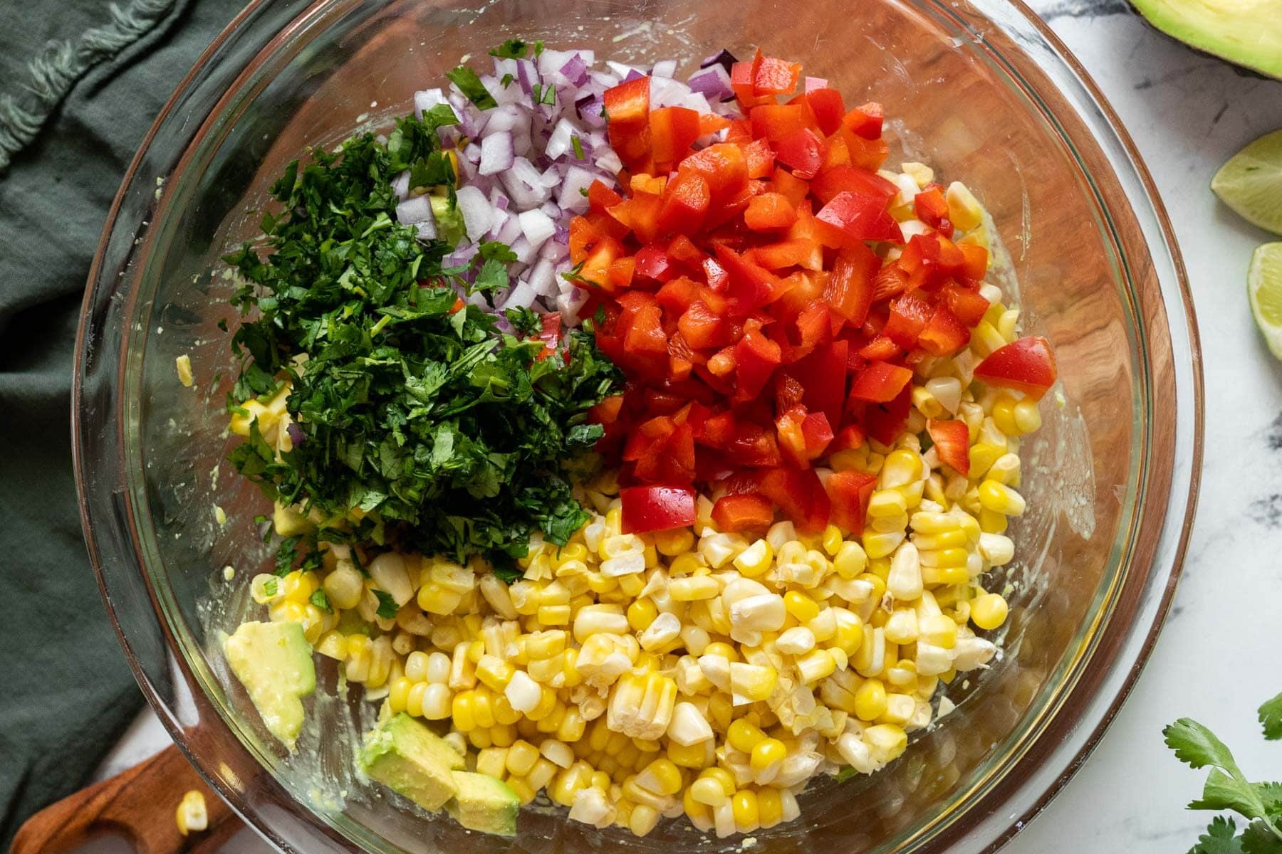 A glass bowl containing chopped cilantro, red onion, red bell pepper, corn, and avocado on a marble surface with lime wedges nearby.