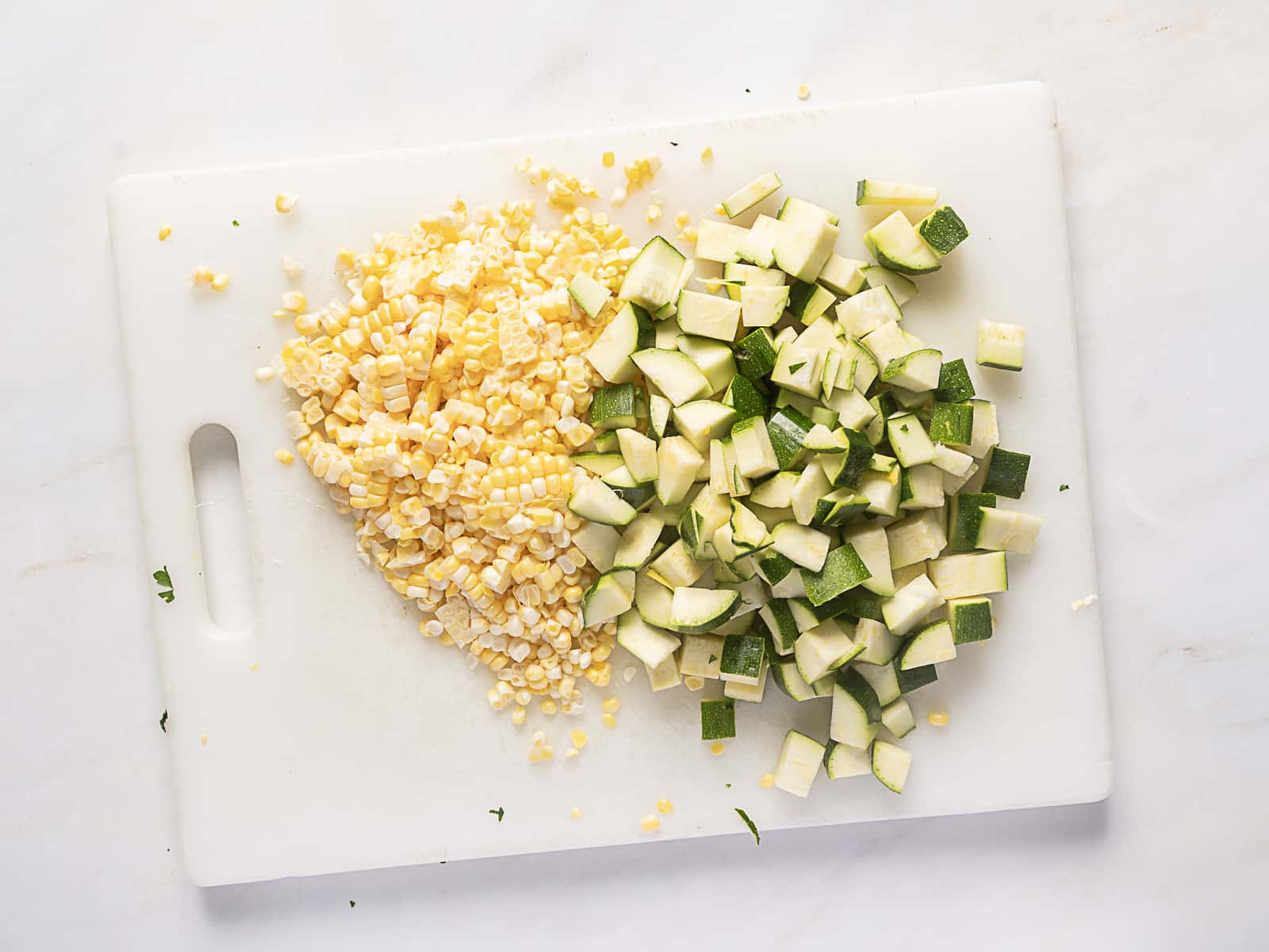 Chopped zucchini and corn kernels are arranged side by side on a white cutting board.