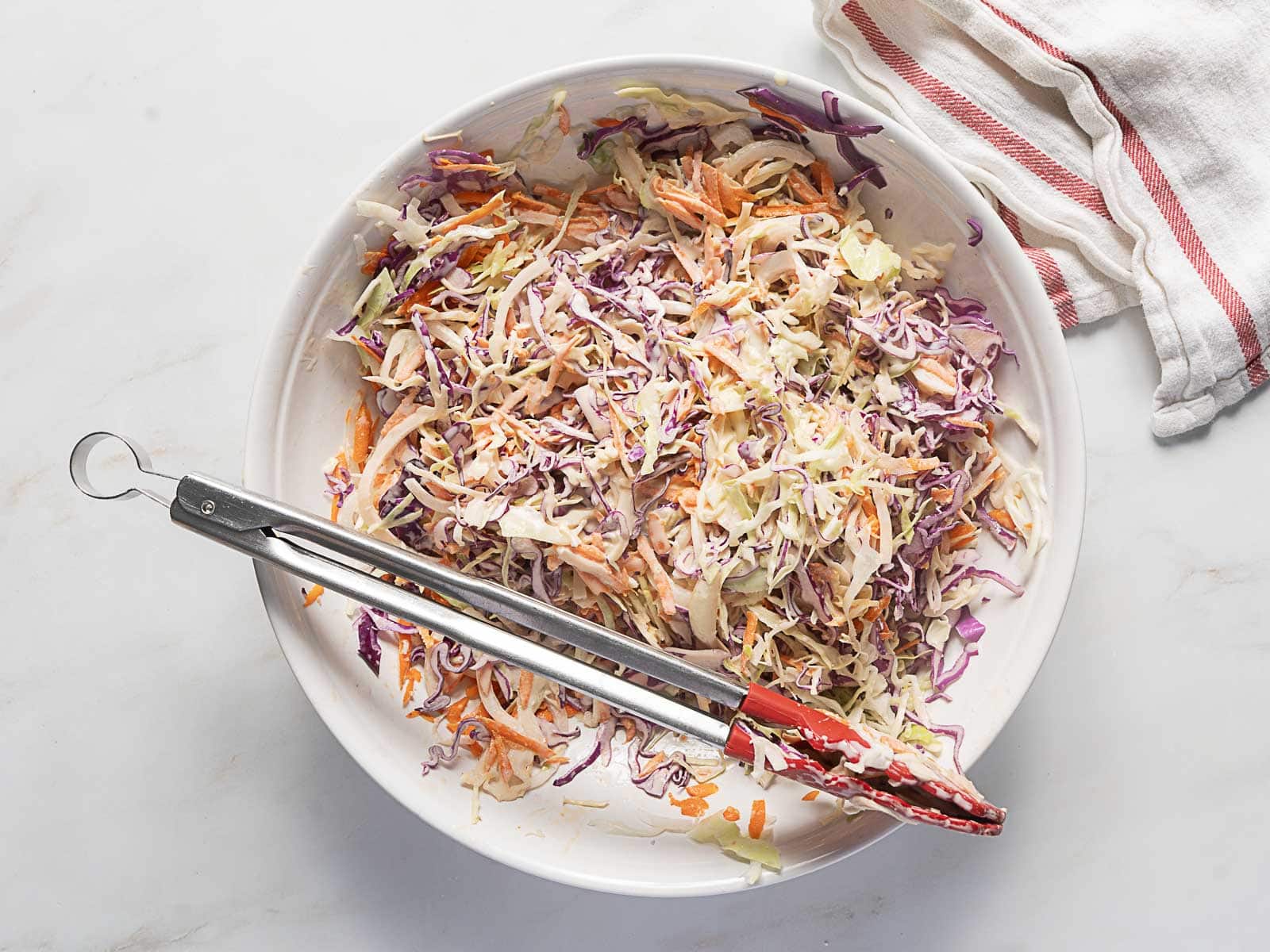 A bowl of coleslaw with shredded cabbage, carrots, and purple cabbage, mixed with dressing, with metal tongs resting on the side.