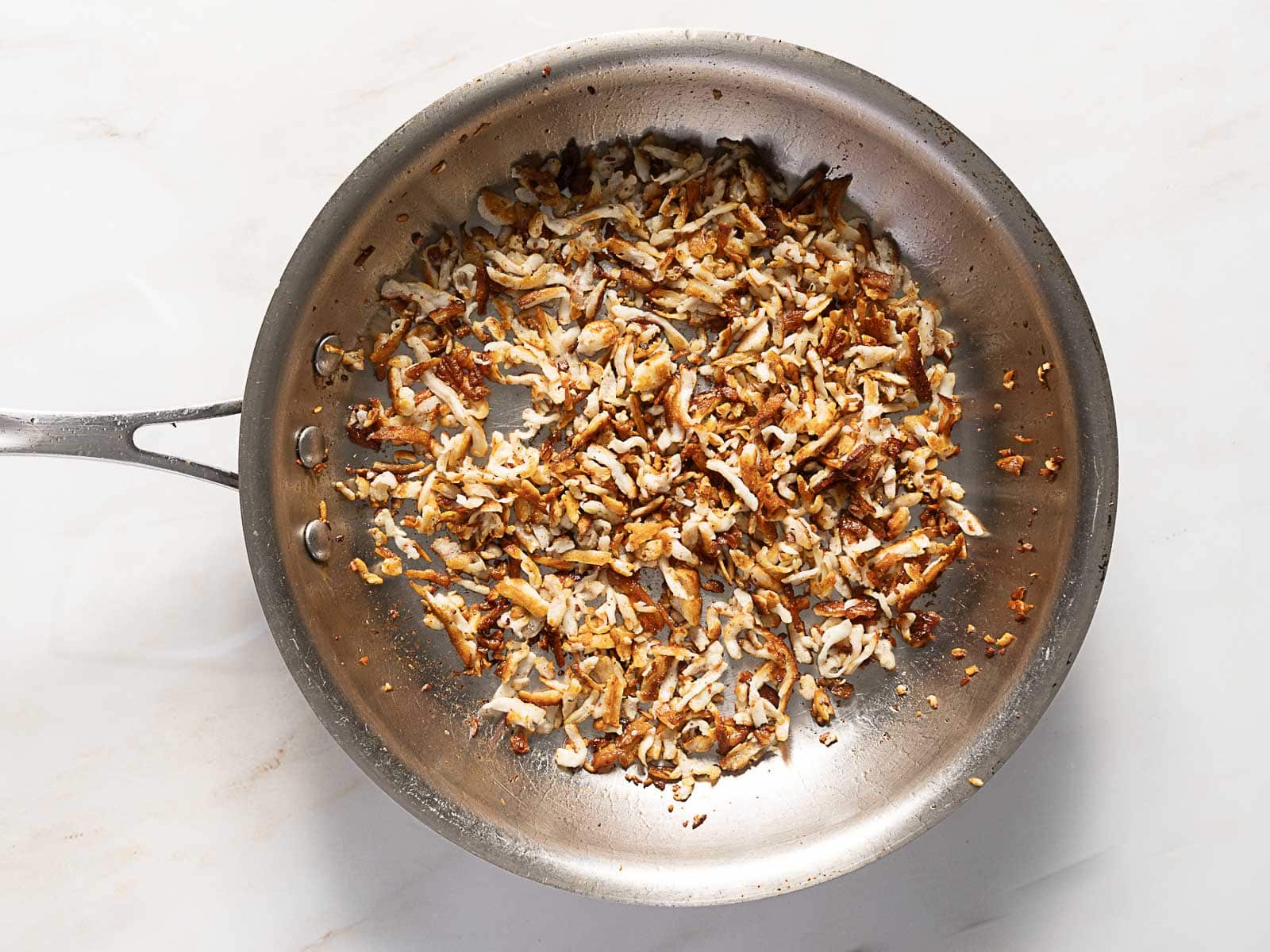 A stainless steel pan containing sautéed and browned shredded potatoes on a light-colored surface.