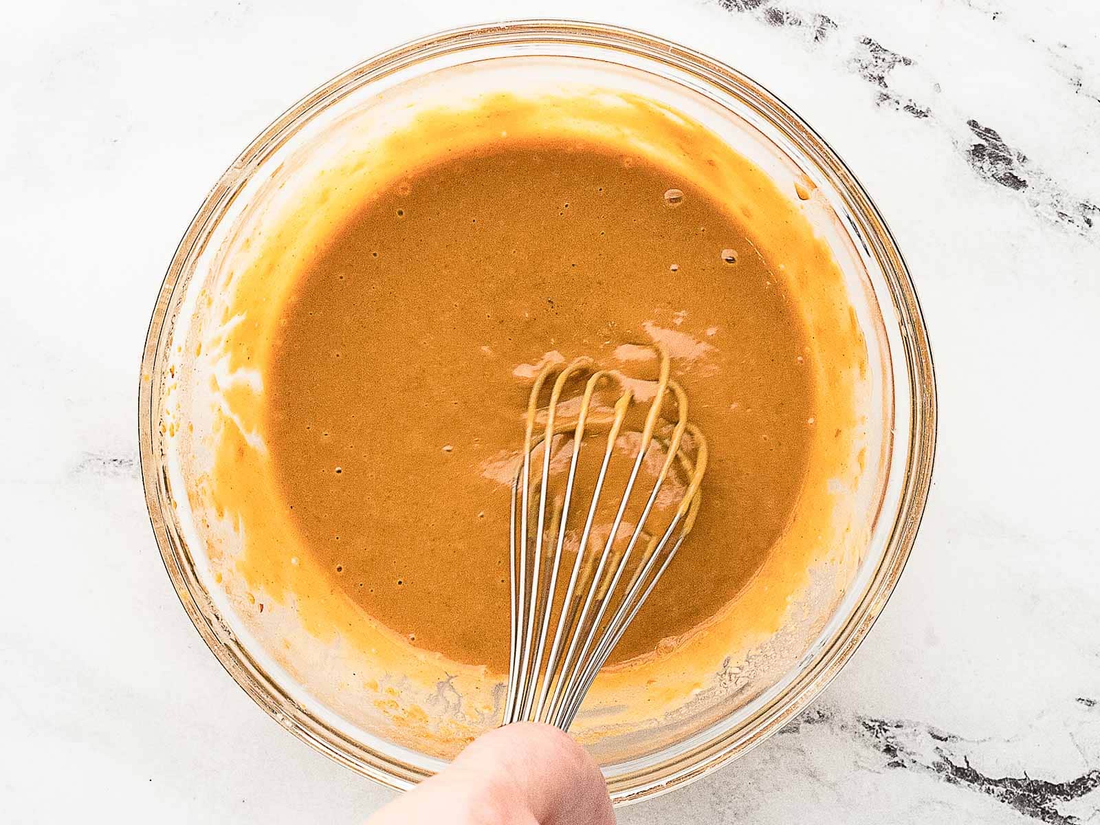 A whisk is mixing a thick, orange-brown sauce in a clear glass bowl on a white marble surface.
