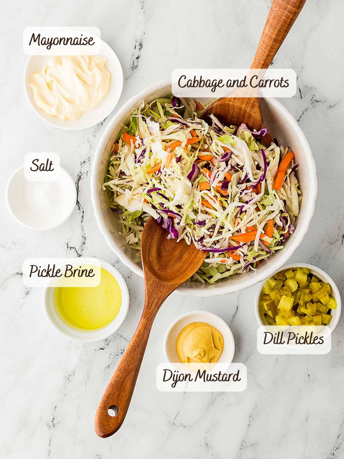 A bowl of shredded cabbage and carrots, perfect for making pickle slaw, with wooden utensils, surrounded by small bowls of mayonnaise, salt, pickle brine, Dijon mustard, and diced dill pickles on a marble surface.