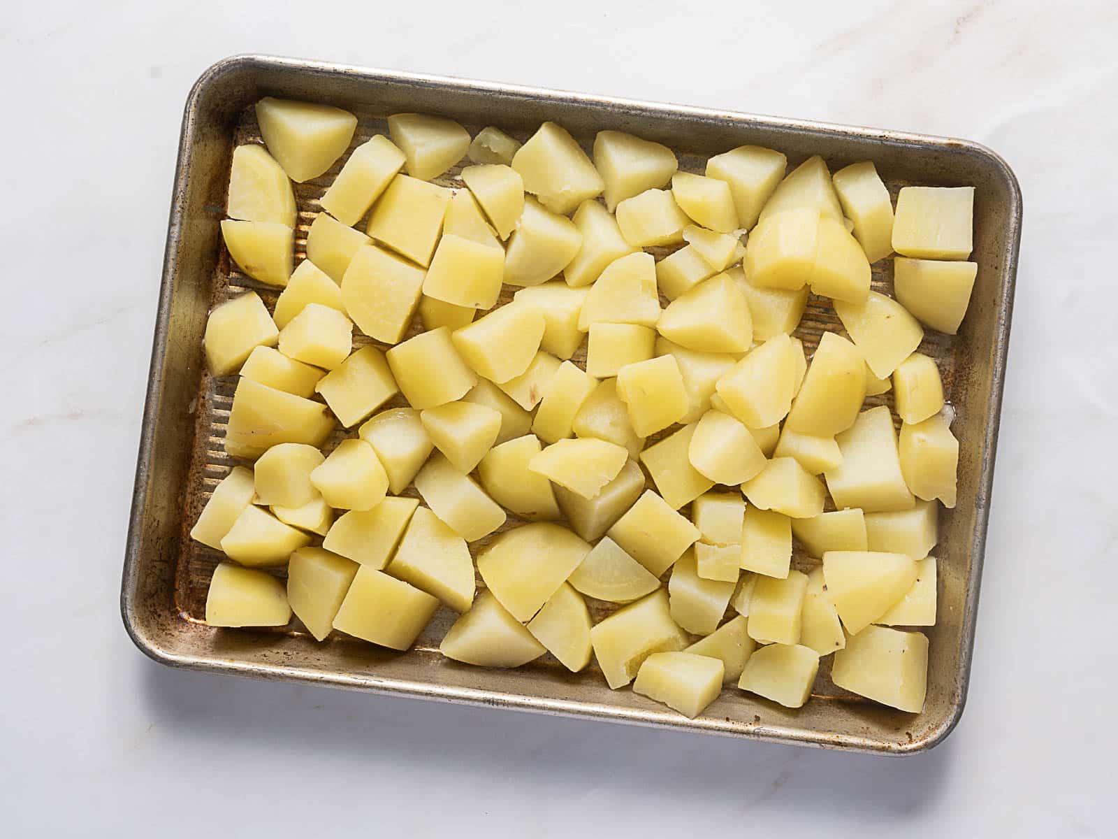 A metal baking sheet filled with evenly spread, peeled, and chopped raw potatoes on a white surface.