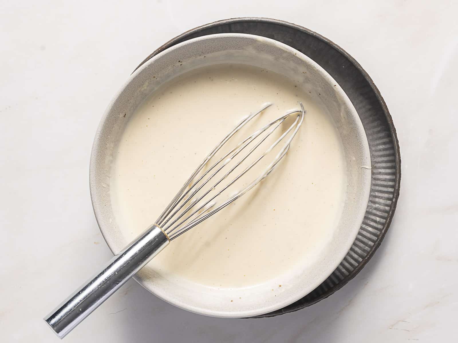 A metal whisk rests in a bowl of creamy white batter, which sits on top of a gray plate on a light-colored surface.