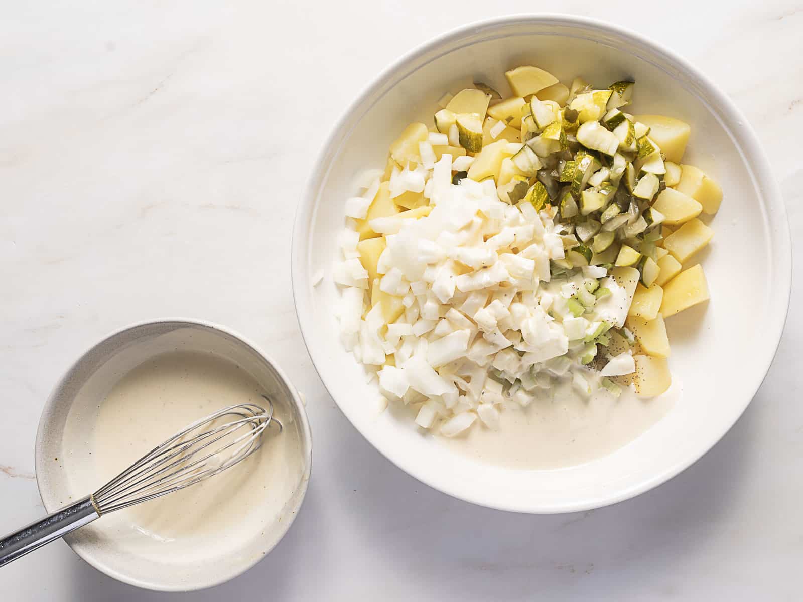 A bowl containing chopped potatoes, eggs, pickles, and celery next to a bowl of whisked creamy dressing on a white surface.