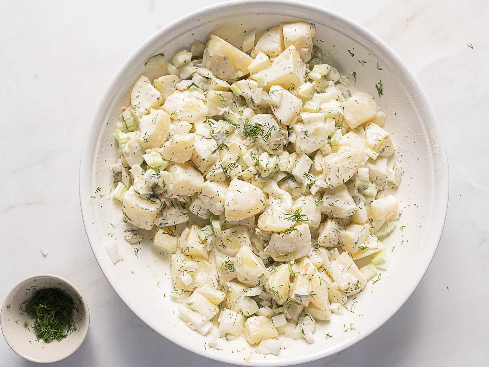 A white bowl filled with potato salad containing chopped potatoes, celery, onions, and fresh dill, with a small dish of extra dill beside it.