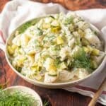 A bowl of creamy potato salad with chopped pickles and fresh dill, set on a wooden surface next to a white cloth and a small dish of dill.