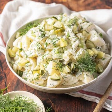 A bowl of creamy potato salad with chopped pickles and fresh dill, set on a wooden surface next to a white cloth and a small dish of dill.