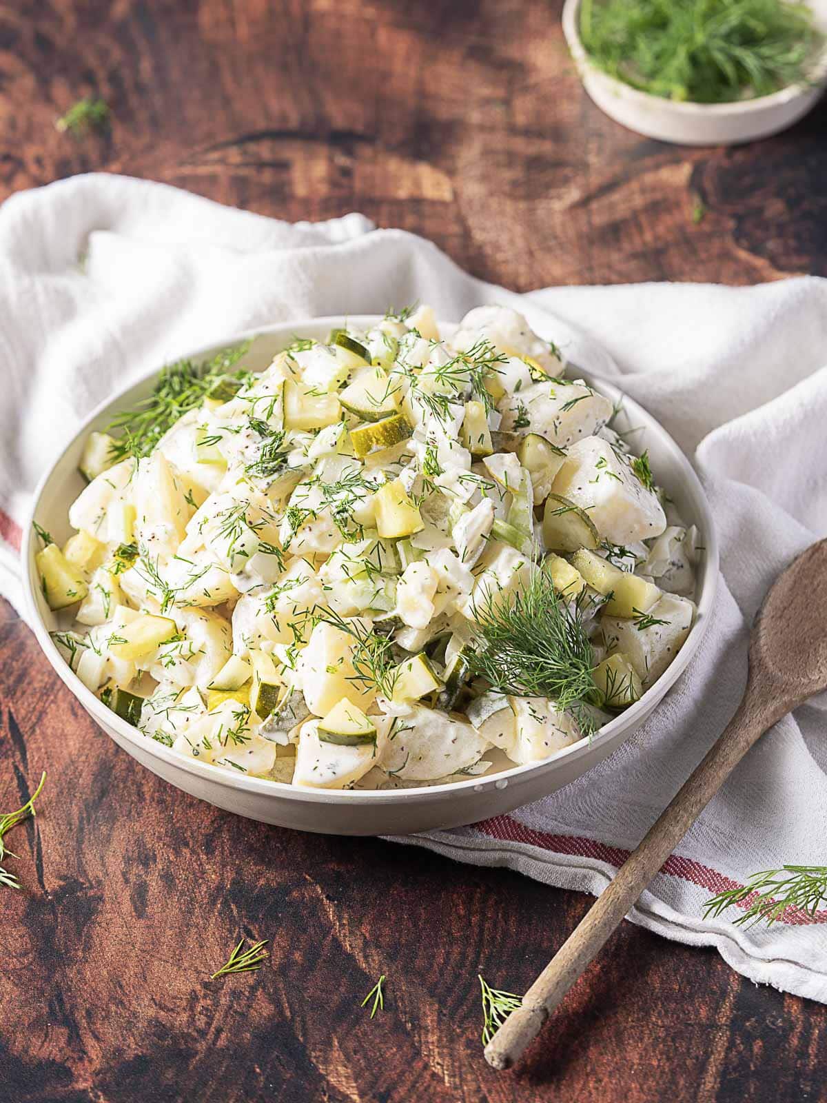 A bowl of creamy potato salad with chopped pickles and fresh dill, placed on a wooden table with a wooden spoon and a white napkin.