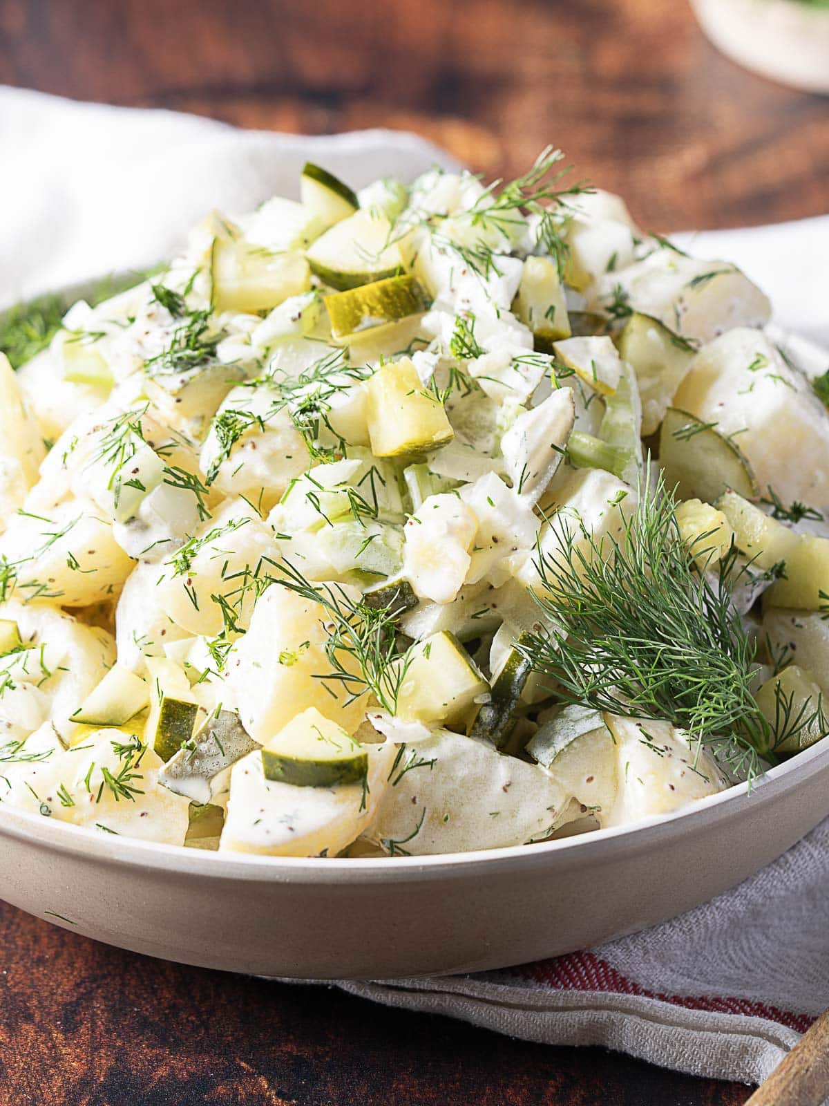 A bowl of potato salad topped with chopped pickles and fresh dill, served on a white plate with a rustic background.