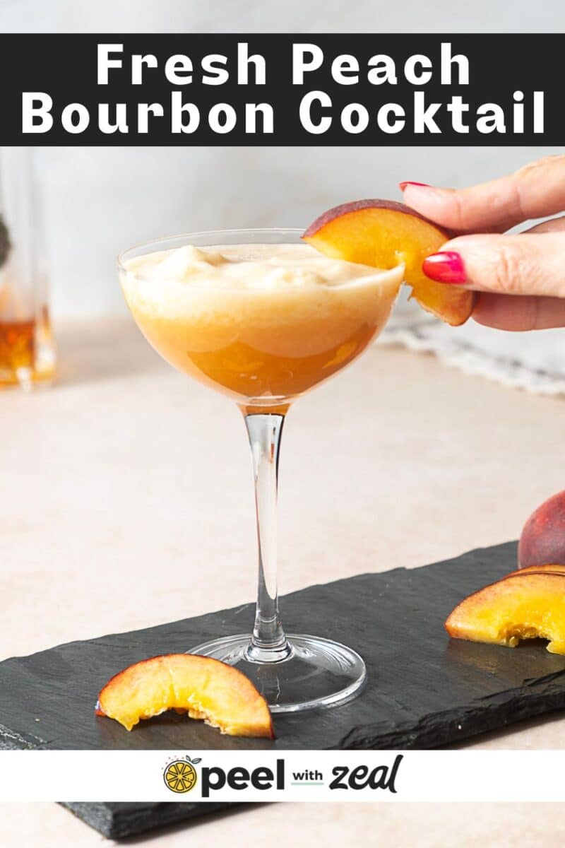 A hand garnishes a coupe glass of peach bourbon cocktail with a fresh peach slice; more peach slices rest on a slate board below.