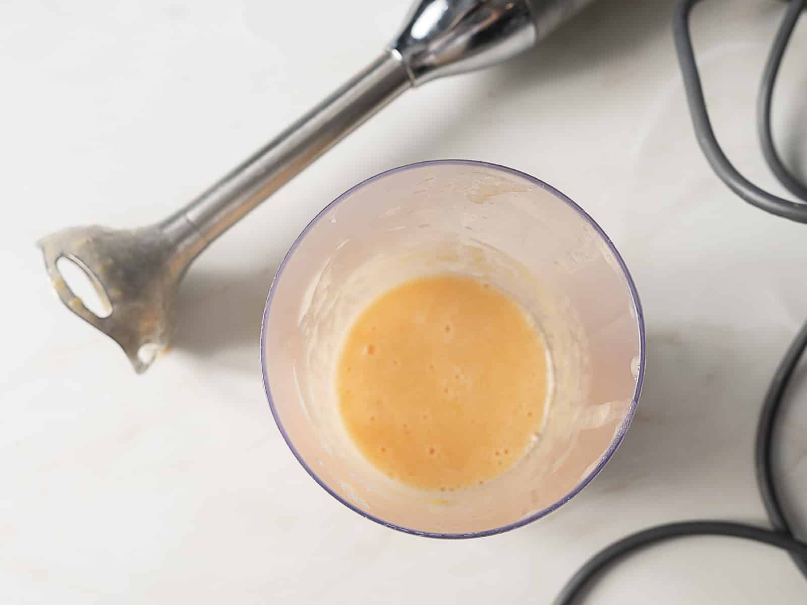 A hand blender and a plastic container with light orange blended mixture are on a white surface.