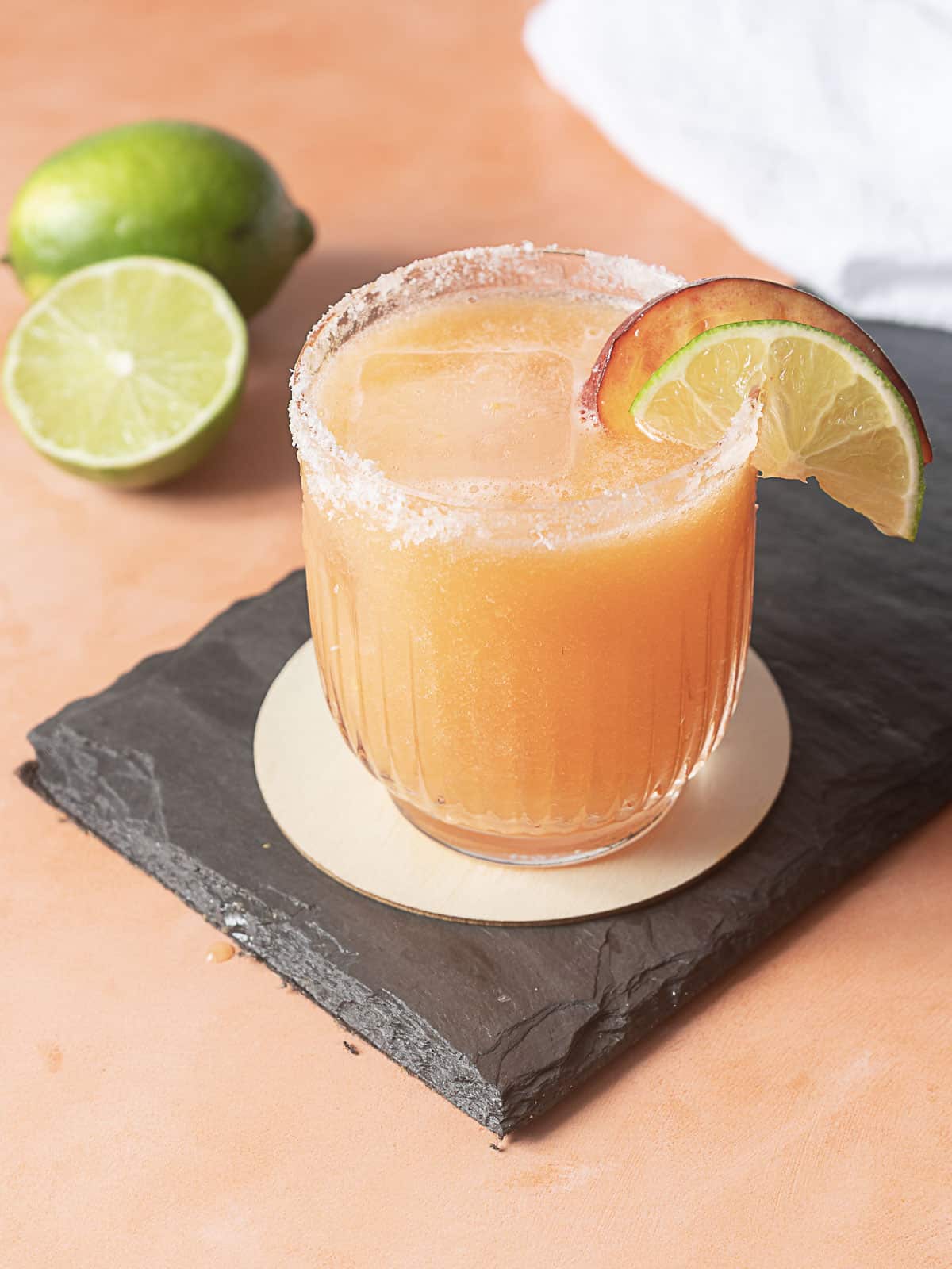 A glass of orange cocktail with a salted rim, garnished with lime and orange slices, sits on a slate coaster. Two limes, one halved, are in the background.