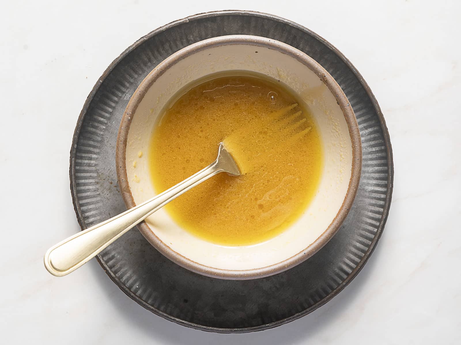 A bowl containing a yellow liquid mixture with a fork resting inside, placed on a dark plate against a white background.