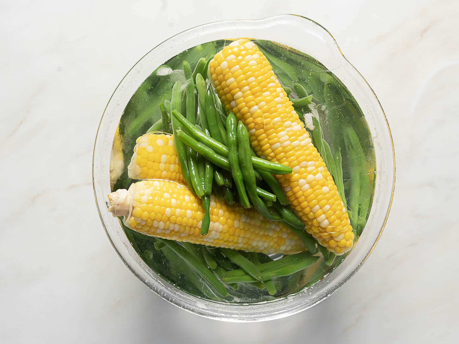 Glass bowl filled with water contains three ears of corn and a handful of green beans on a white surface.