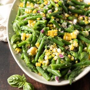 A platter of green bean and corn salad with chopped red onion, fresh dill, and basil leaves, served on a white oval dish.