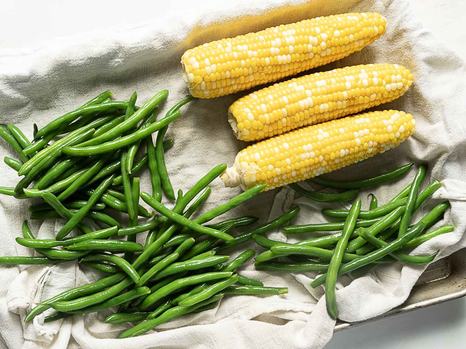 Three ears of corn and a pile of fresh green beans are arranged on a cloth-covered surface.