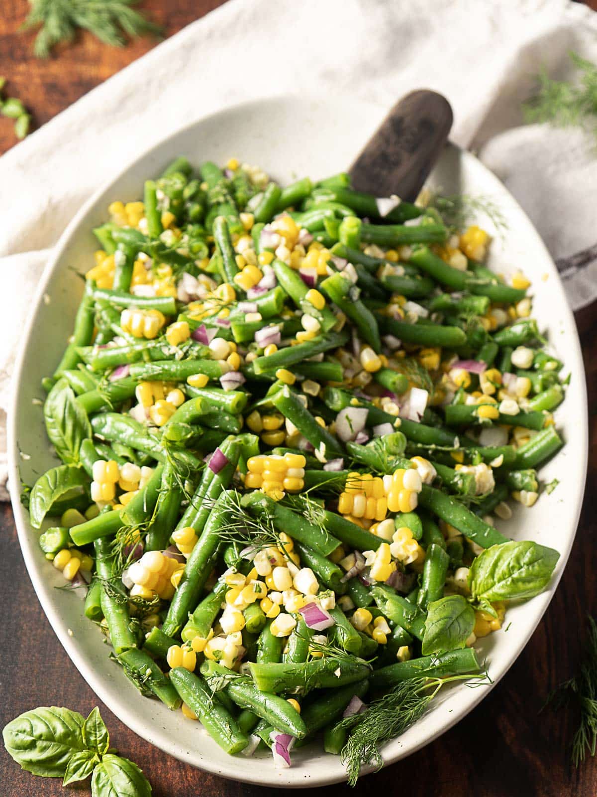 A white oval plate filled with a salad of green beans, corn kernels, chopped red onion, and fresh herbs, with a serving spoon on the side.