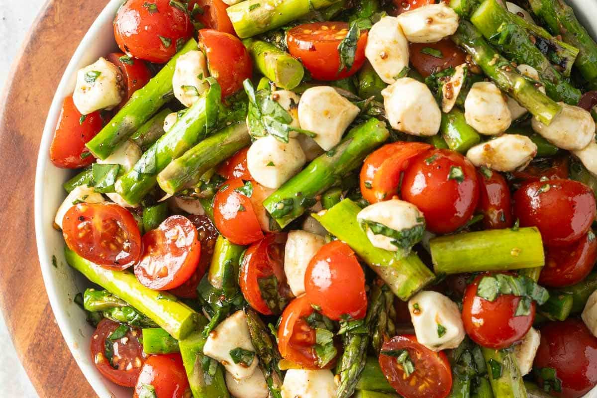 A bowl of salad with cherry tomatoes, asparagus, mozzarella balls, chopped basil, and a vinaigrette dressing.