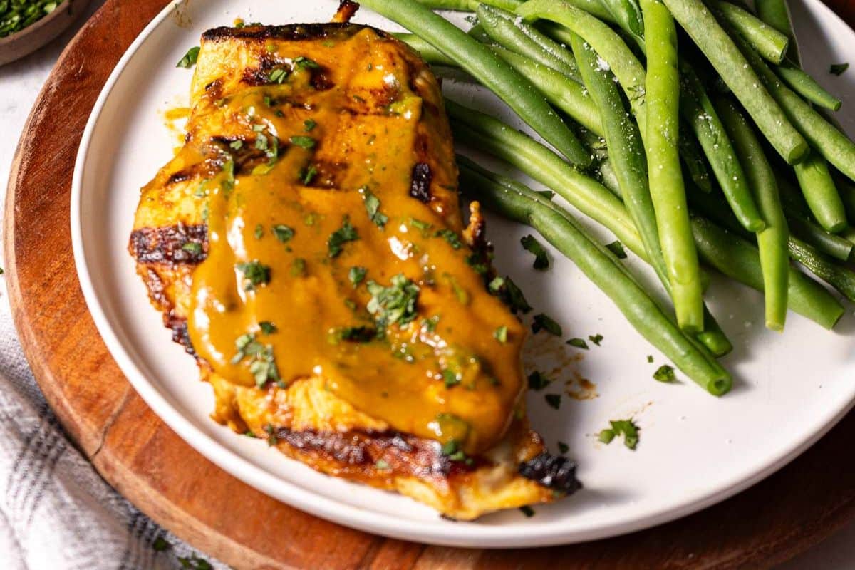 A plate with grilled chicken covered in yellow sauce, garnished with herbs, served alongside steamed green beans.