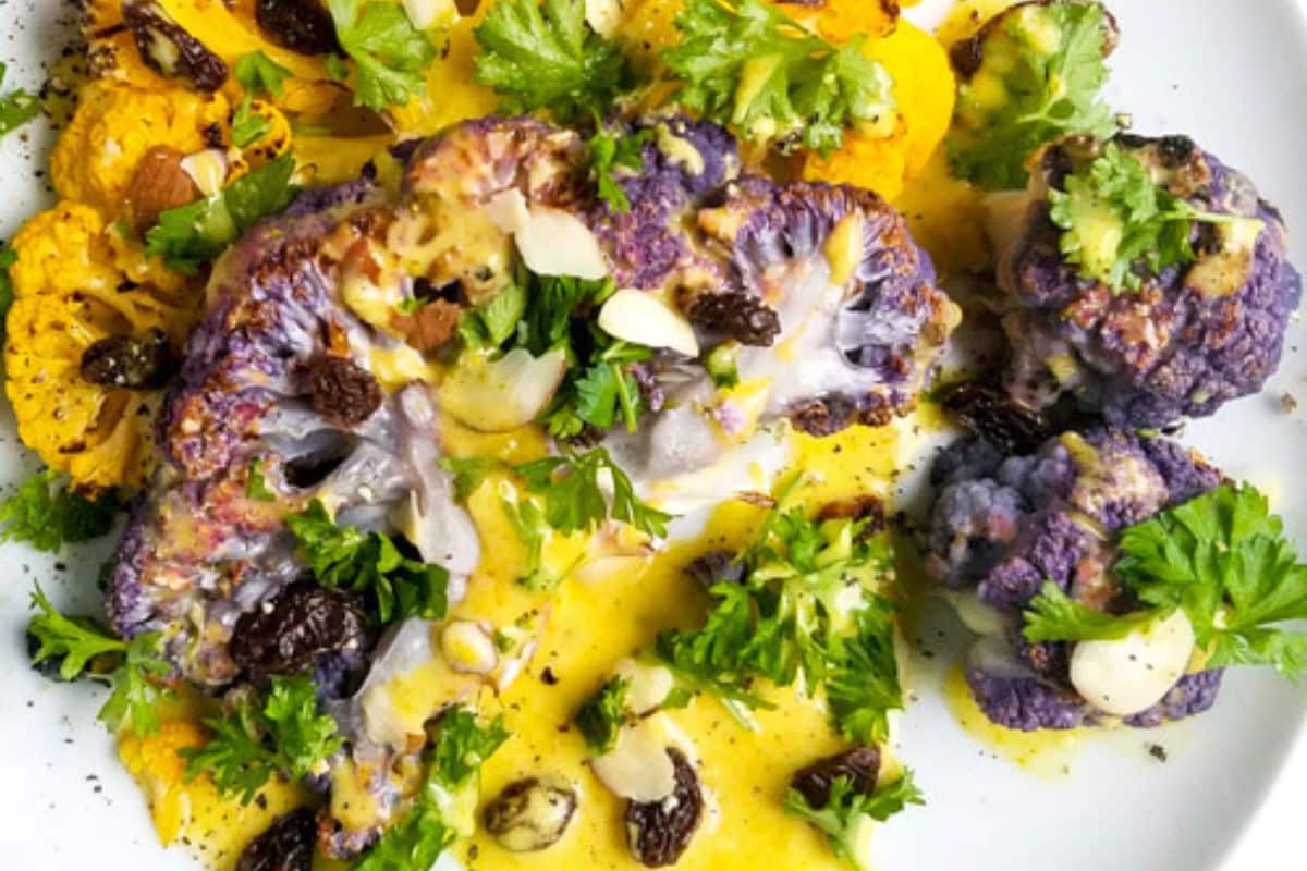 Roasted yellow and purple cauliflower on a white plate, topped with sauce, raisins, sliced almonds, and fresh parsley.