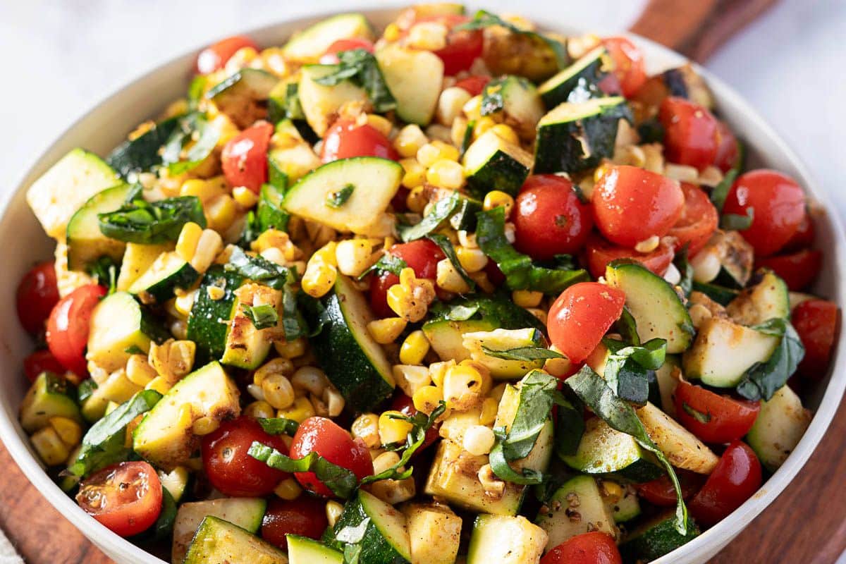 A bowl of fresh salad containing chopped zucchini, cherry tomatoes, corn, and basil, lightly dressed, served on a wooden board.