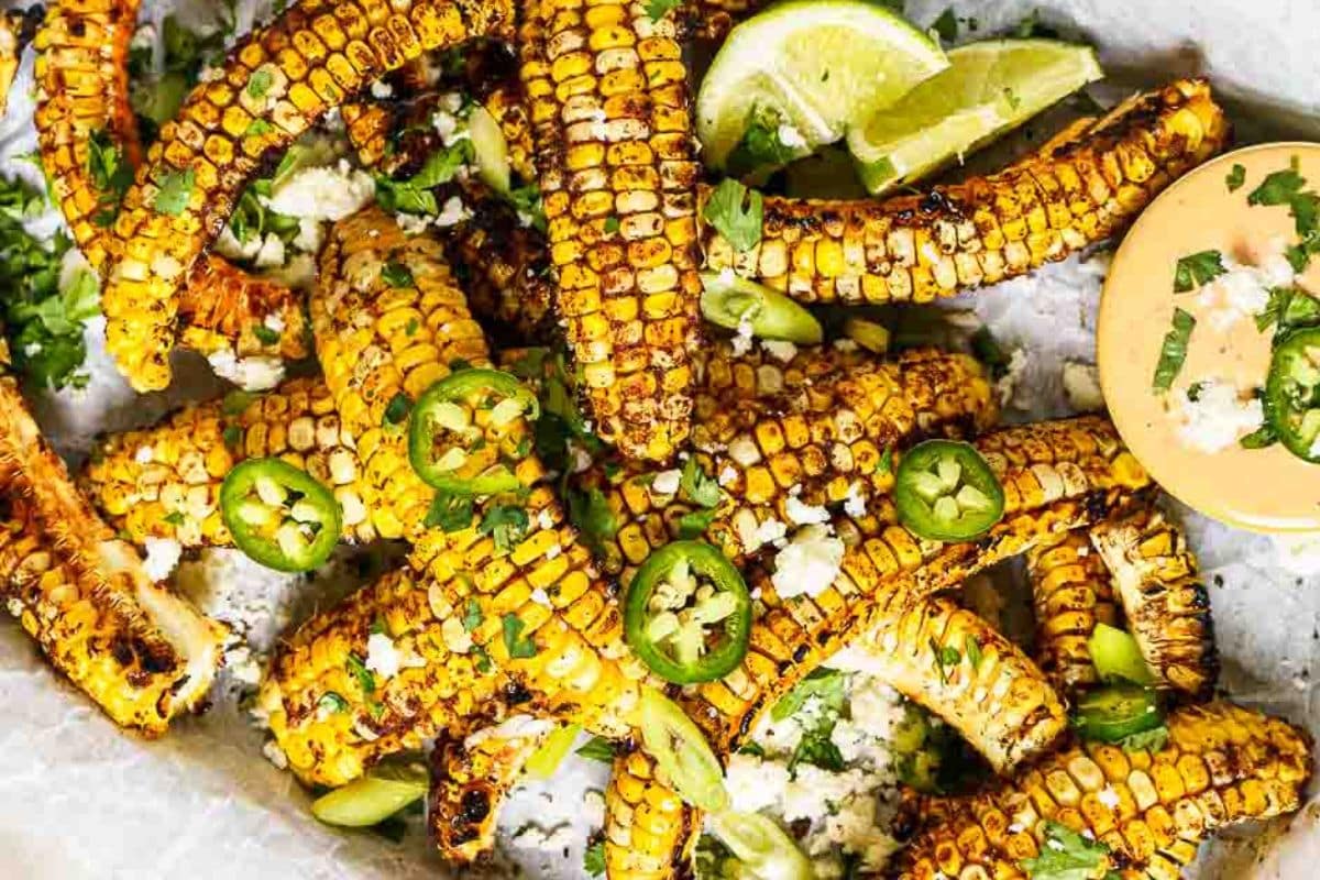 Grilled corn ribs topped with cheese, sliced jalapeños, cilantro, and green onions, served with lime wedges and a creamy dipping sauce.