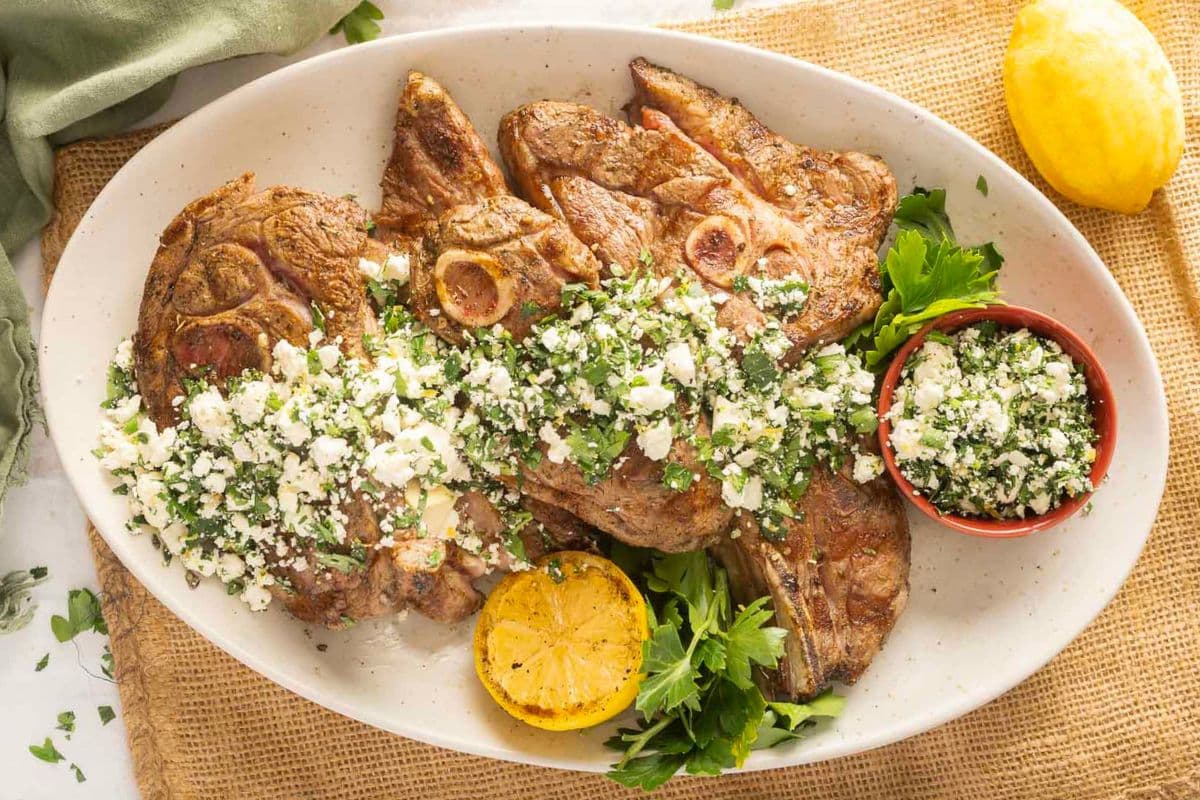 Grilled lamb chops topped with a feta and herb mixture, served on a white platter with grilled lemon halves and fresh parsley garnish.