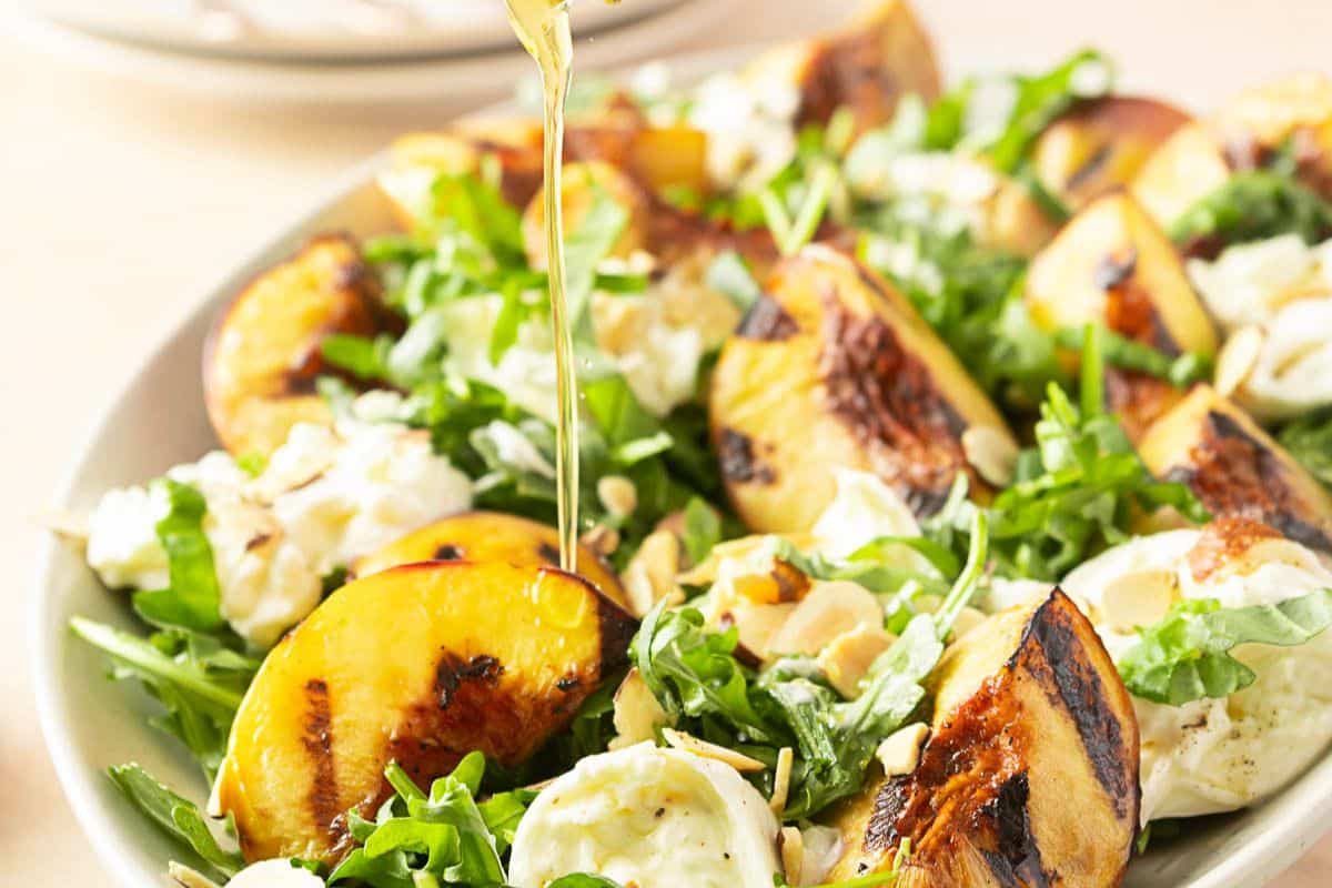 A plate of salad with grilled peach slices, arugula, mozzarella, and nuts being drizzled with dressing.