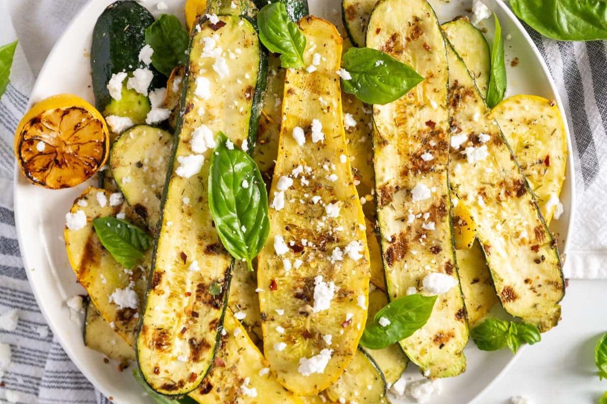 Sliced roasted zucchini and yellow squash on a plate, garnished with crumbled cheese, basil leaves, and a grilled lemon half.