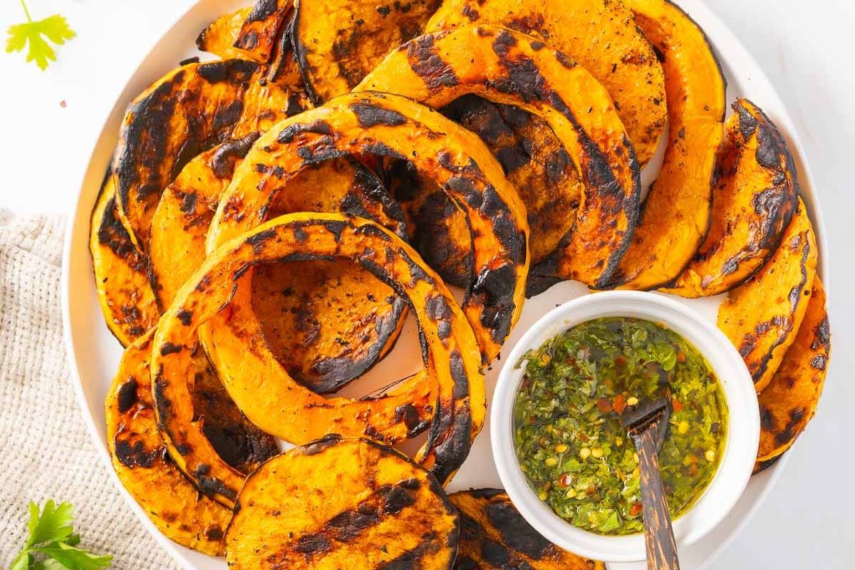 Grilled slices of squash arranged on a white plate, served with a small bowl of green herb sauce and a spoon.