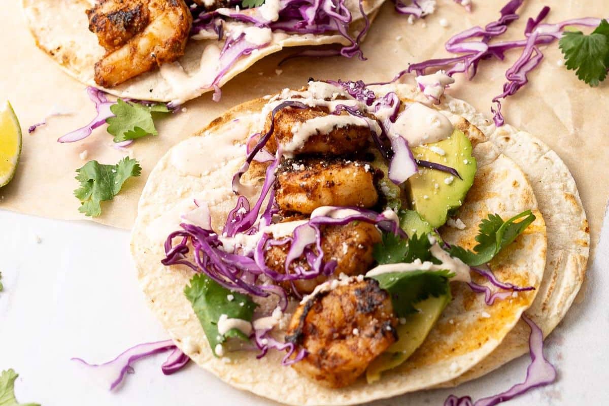 Two shrimp tacos on corn tortillas topped with grilled shrimp, sliced avocado, shredded red cabbage, fresh cilantro, and a drizzle of creamy sauce.