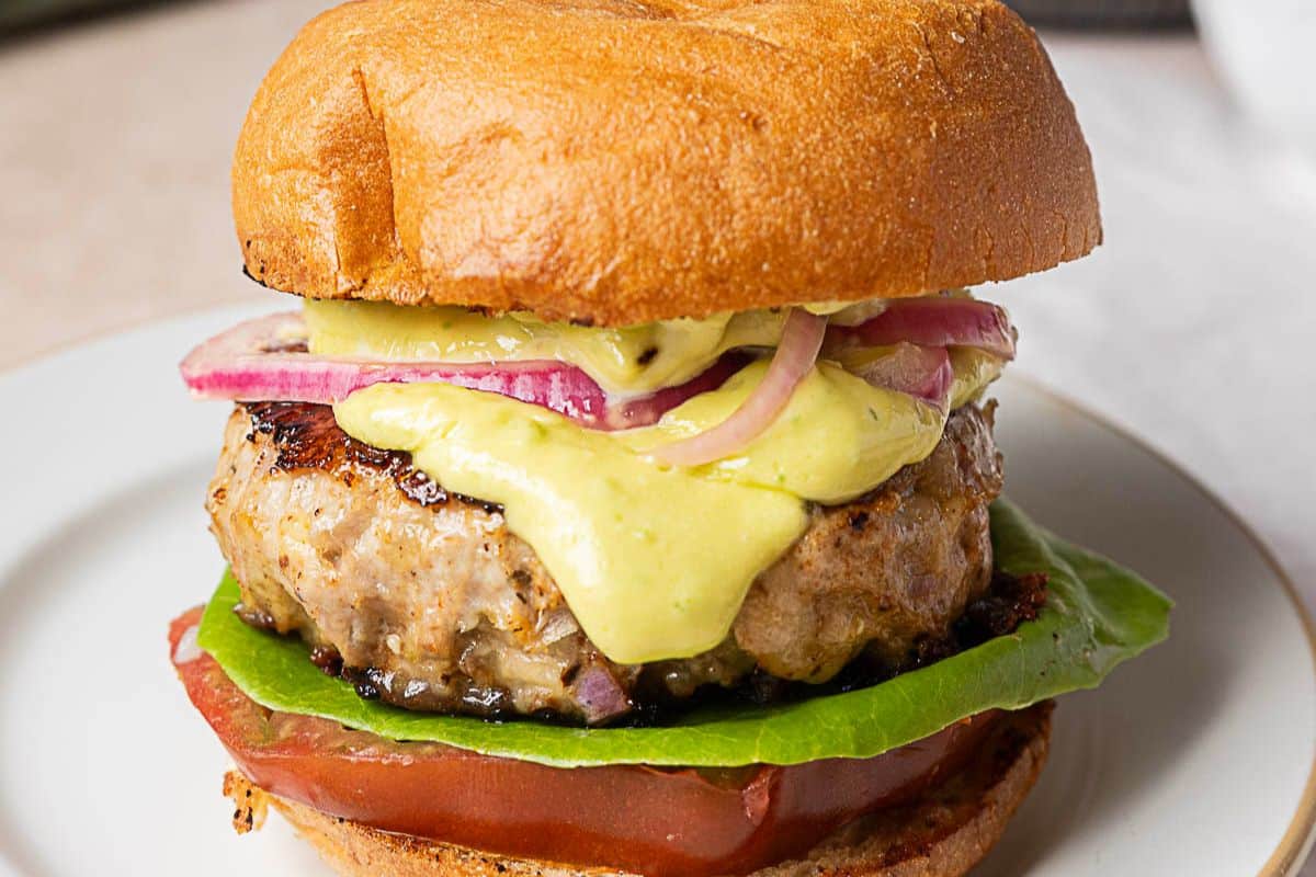 A close-up of a sandwich with a grilled patty, lettuce, tomato, red onions, and yellow sauce on a toasted bun, served on a white plate.