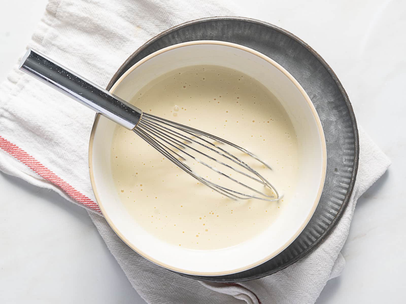 A metal whisk rests in a bowl of pale batter on a gray plate, placed on a folded white kitchen towel with a red stripe.