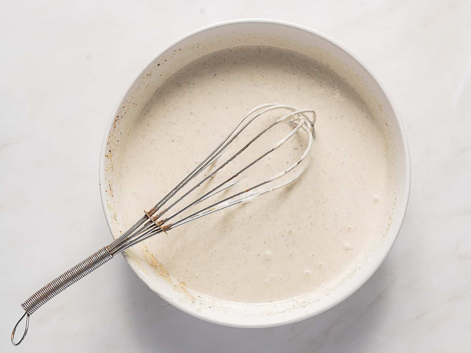 A metal whisk rests in a bowl of smooth, light brown batter on a white surface.