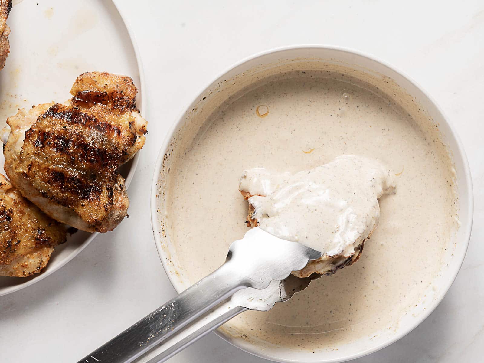 Grilled chicken is held with tongs and dipped into a bowl of creamy white sauce, with more grilled chicken on a plate nearby.