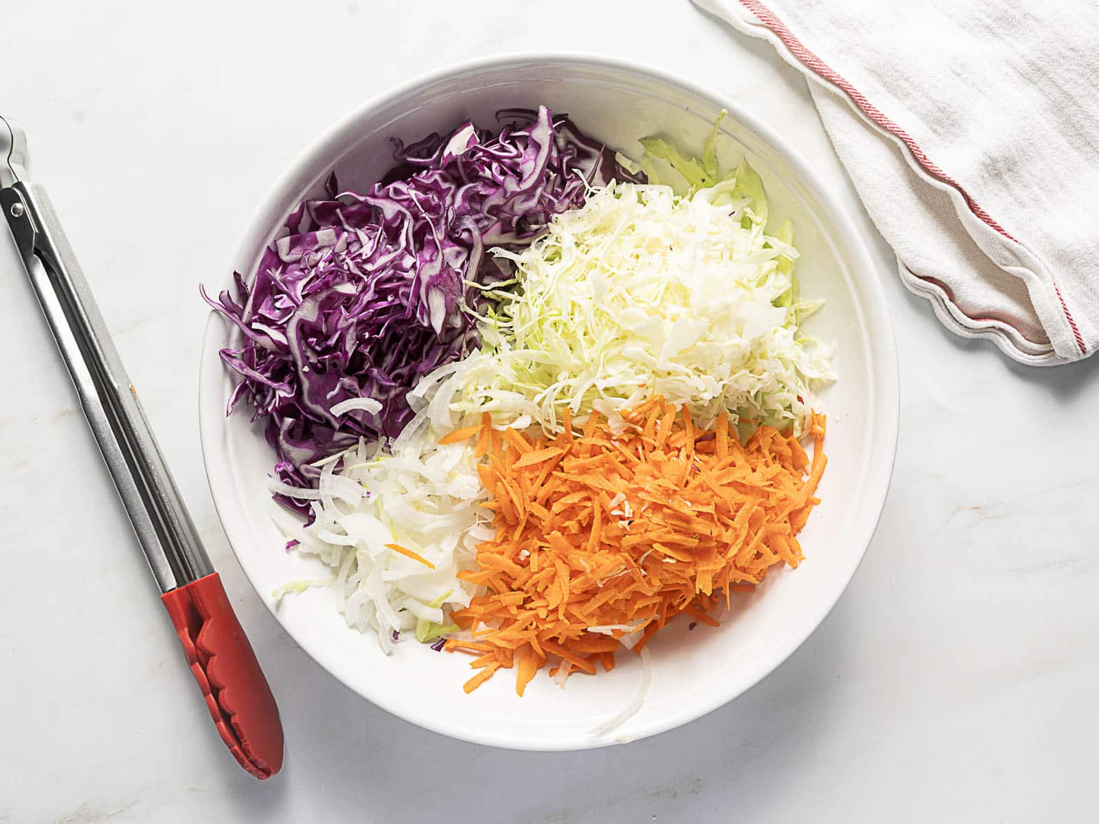 A white bowl contains shredded red cabbage, green cabbage, and carrots, placed on a white surface next to metal tongs and a folded white towel.