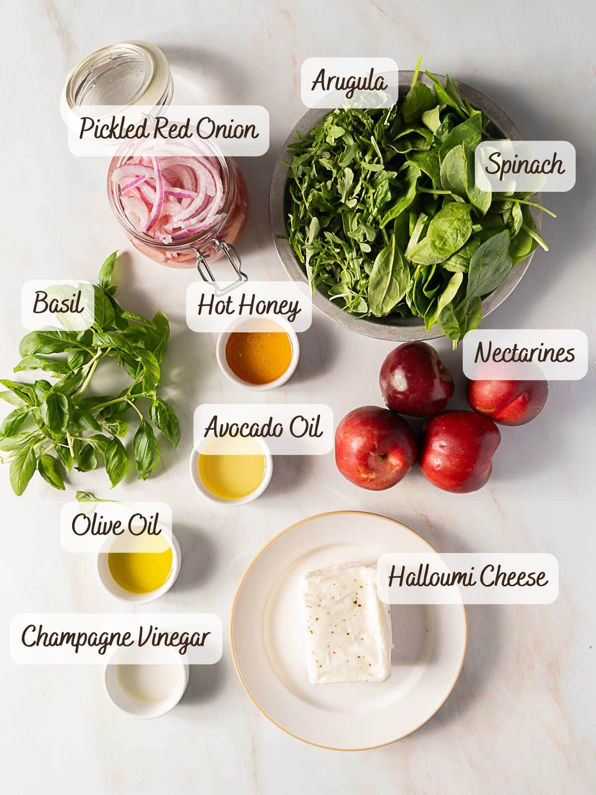 Ingredients for a nectarine salad arranged on a white surface: pickled red onion, arugula, spinach, basil, nectarines, halloumi cheese, avocado oil, olive oil, champagne vinegar, and hot honey.