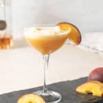 A coupe glass filled with a peach-colored cocktail, garnished with a peach slice on the rim, sits on a slate surface with additional peach slices nearby.