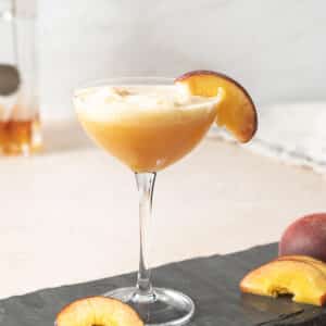 A coupe glass filled with a peach-colored cocktail, garnished with a peach slice on the rim, sits on a slate surface with additional peach slices nearby.